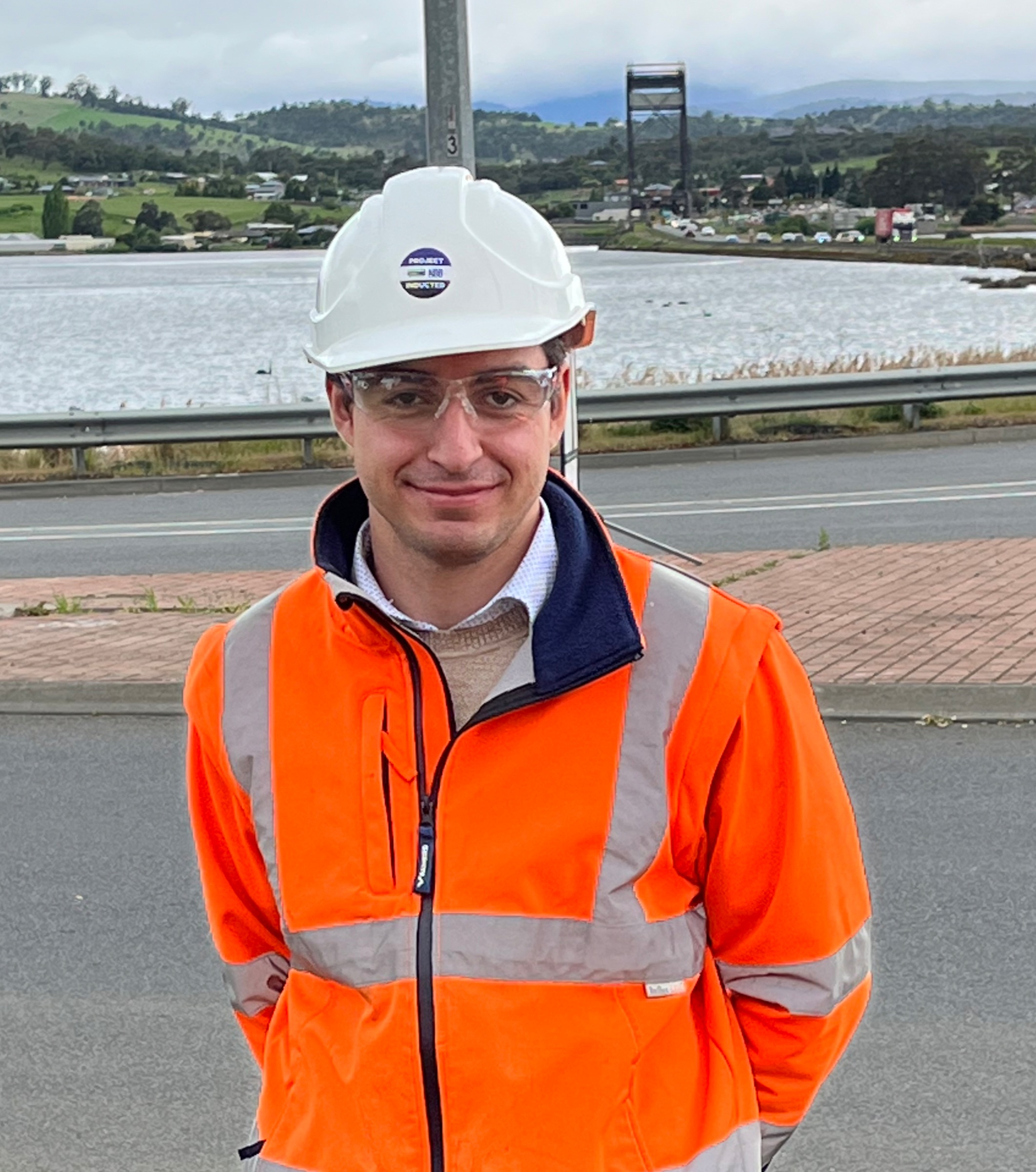 A former TasTAFE student Kosta Vasileiou in Hi-Vis clothing wiht a road and water behind him 