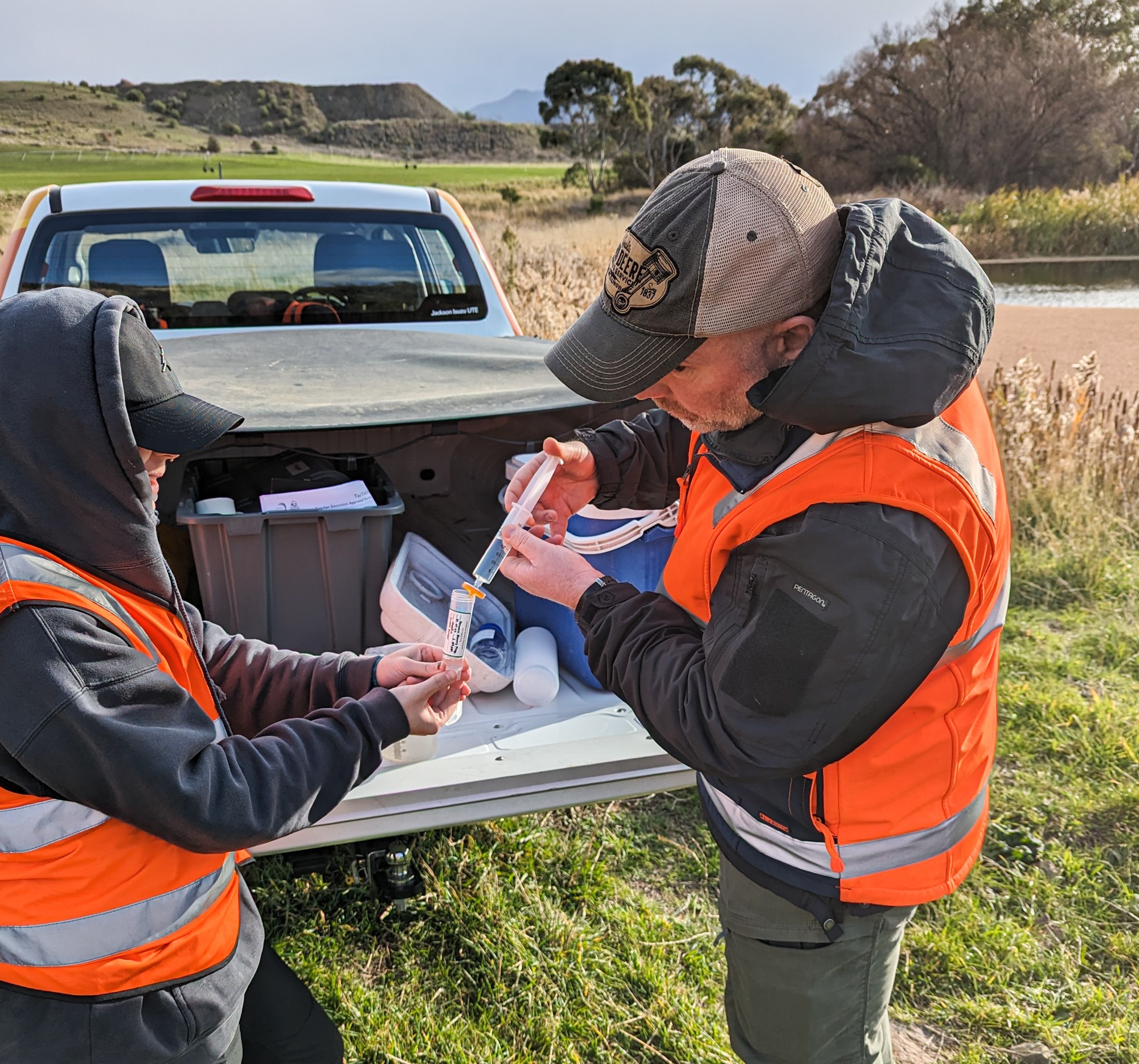 Two people wearing Hi Visibility clothing using a water syringe in front of a ute at an estuary